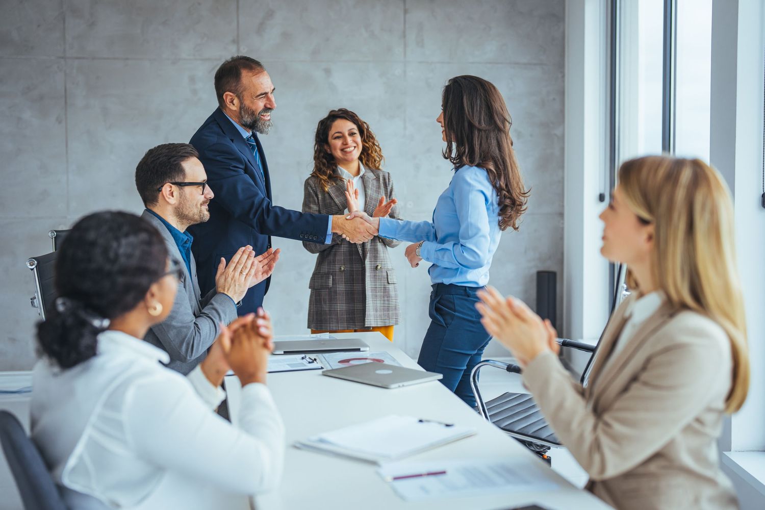 Business professionals shaking hands across a conference table while colleagues applaud in a modern office meeting room.