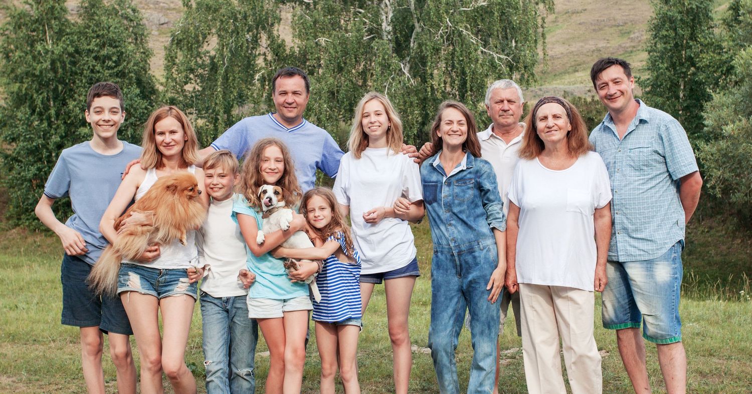 Multi-generational family with two dogs smiling outdoors on a sunny day with lush green trees in the background.