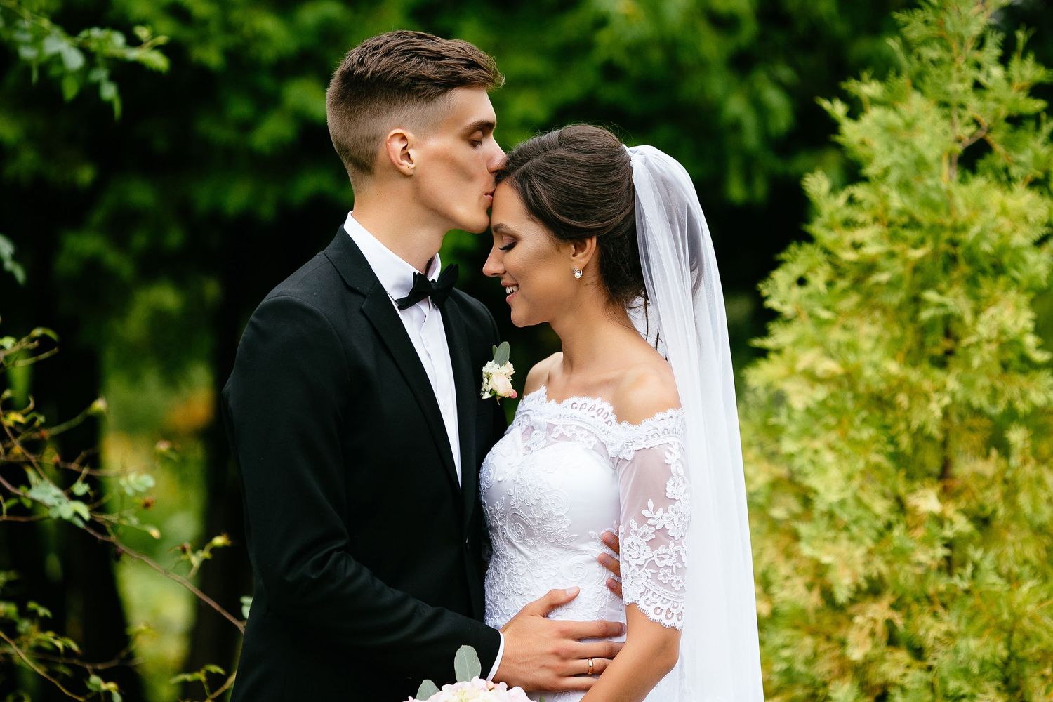Bride and groom share a romantic kiss outdoors surrounded by lush green trees in elegant wedding attire.