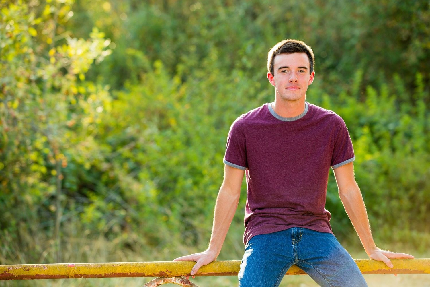 Young man in purple t-shirt and jeans leaning on yellow railing outdoors with green trees in background.