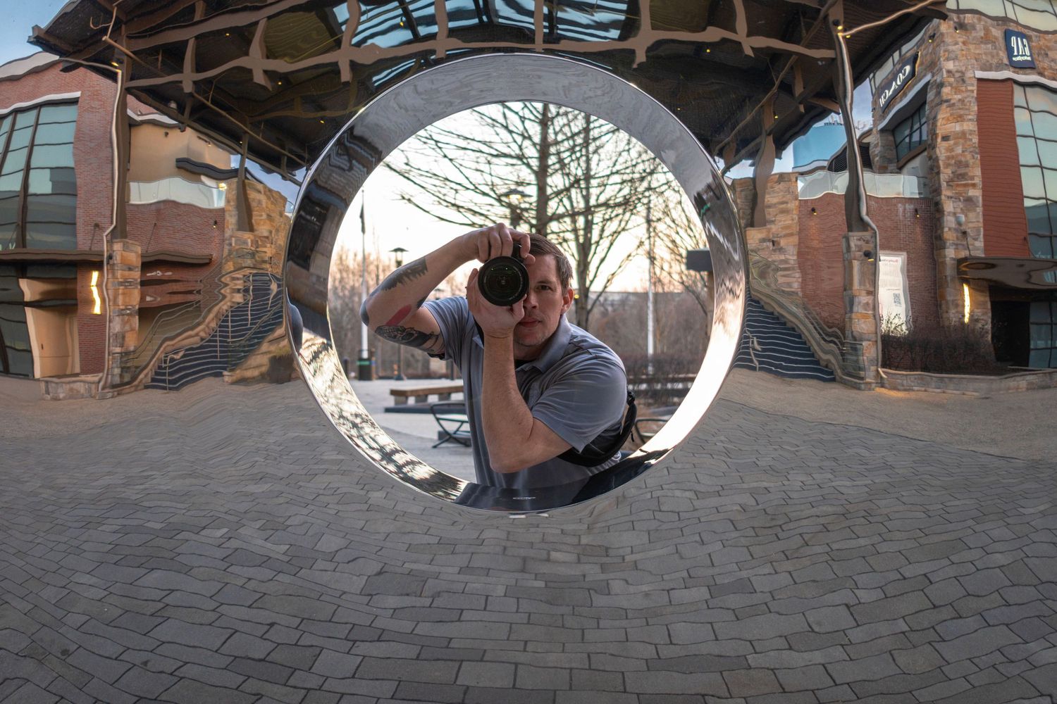 Photographer captures self-portrait reflected in circular mirror sculpture outdoors, fish-eye lens distortion visible.