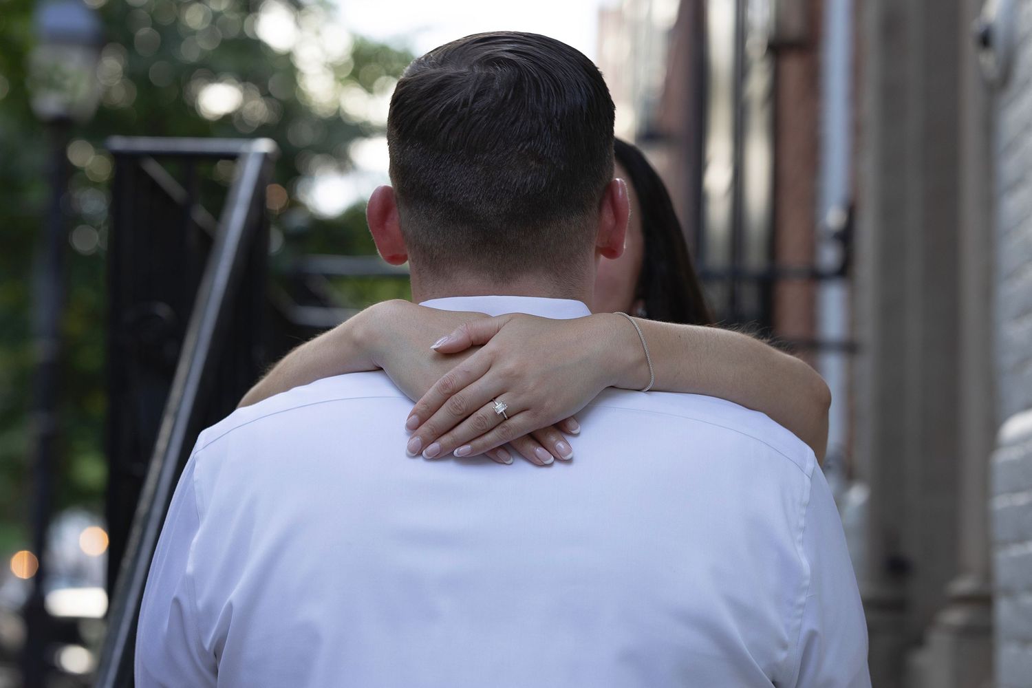 A couple embraces on a city street, woman wearing an engagement ring with her arm around mans neck.