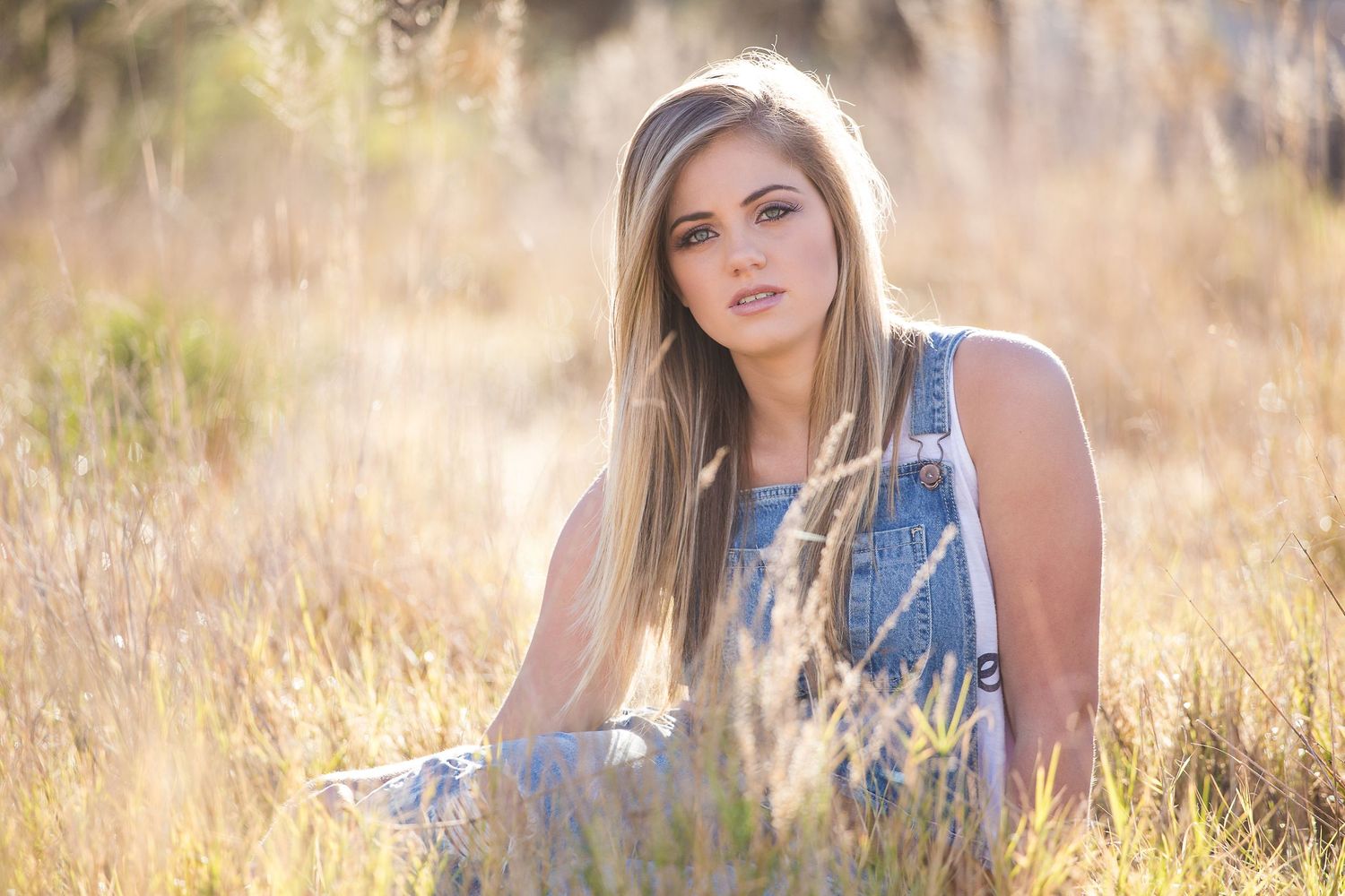 Young blonde woman in denim overalls sitting in golden grass field, bathed in warm sunlight.