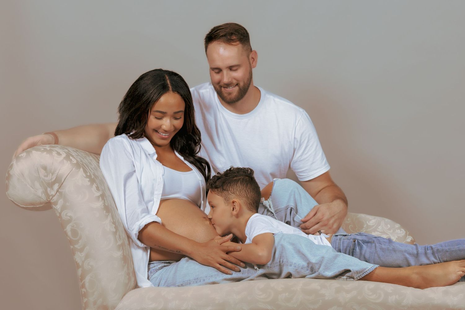 Pregnant woman in white shirt with husband and young son kissing her baby bump on a cozy chair.