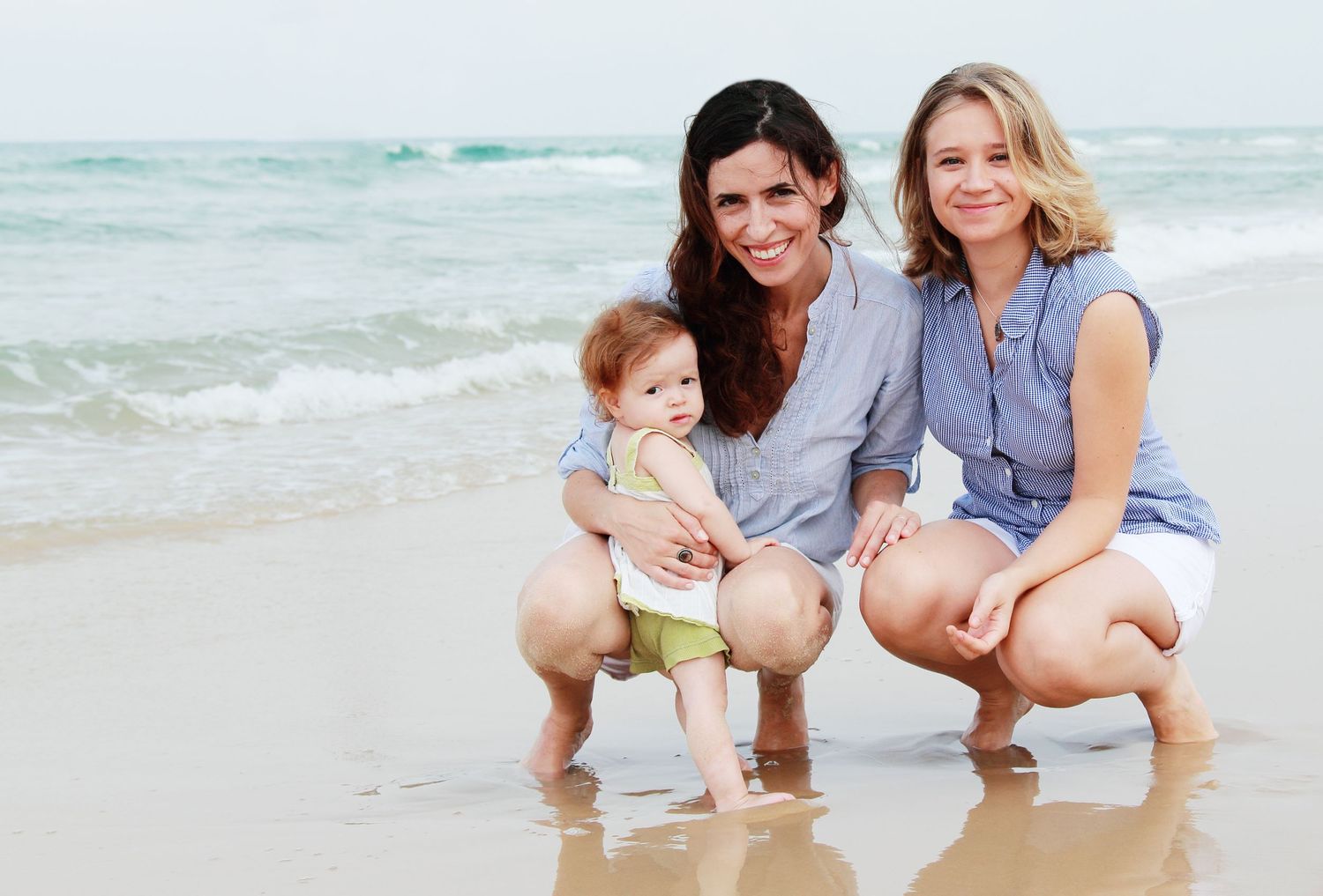 Two women and a baby posing together on a sandy beach with ocean waves in the background.