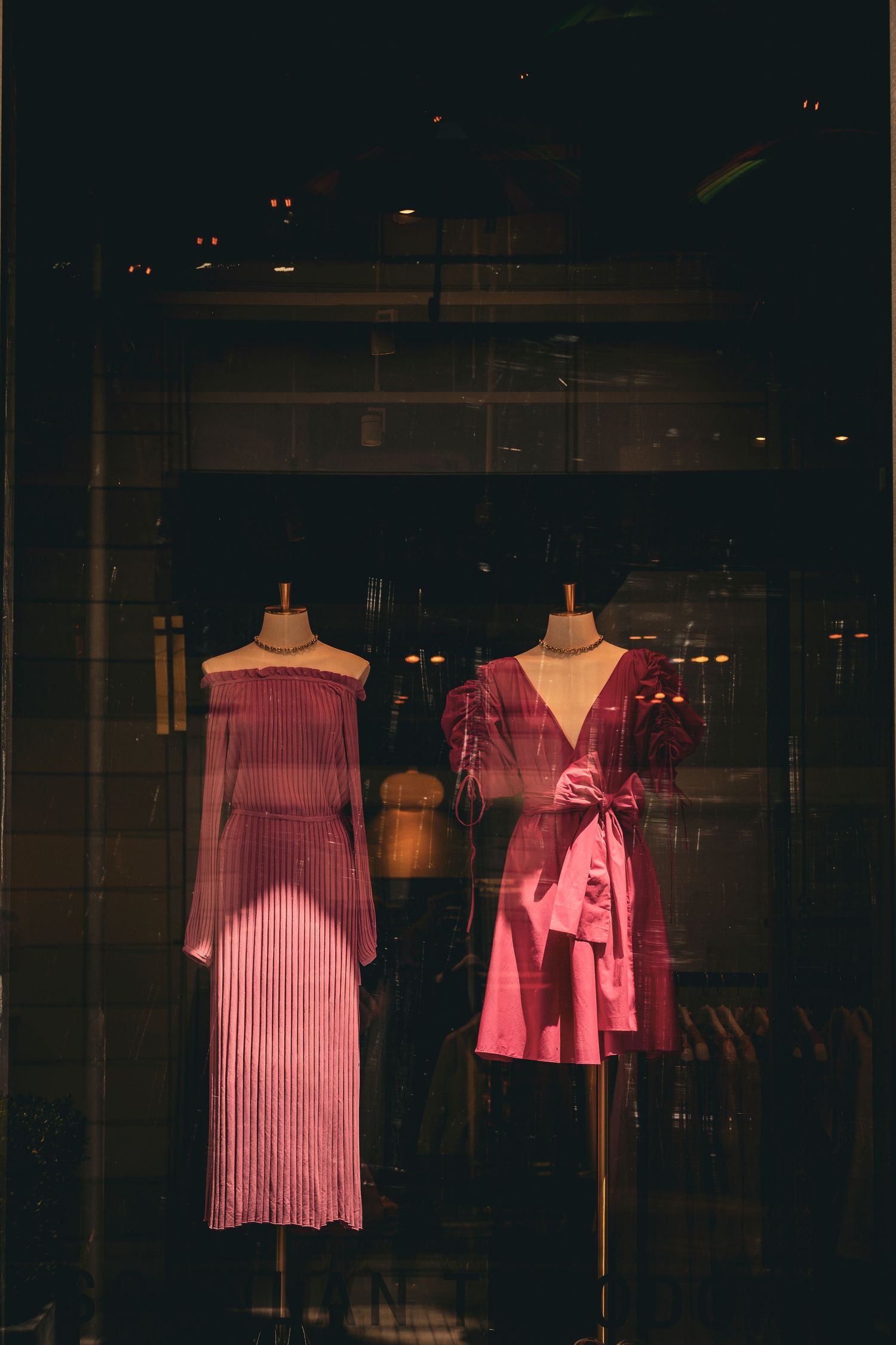 Two pink dresses on mannequins displayed in a dimly lit boutique store window at night.