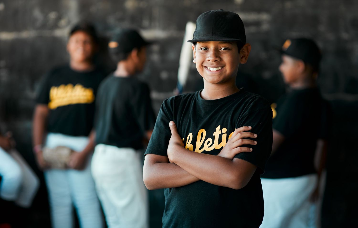 Young boy in Athletics baseball uniform smiles confidently with arms crossed in dugout with teammates behind him.