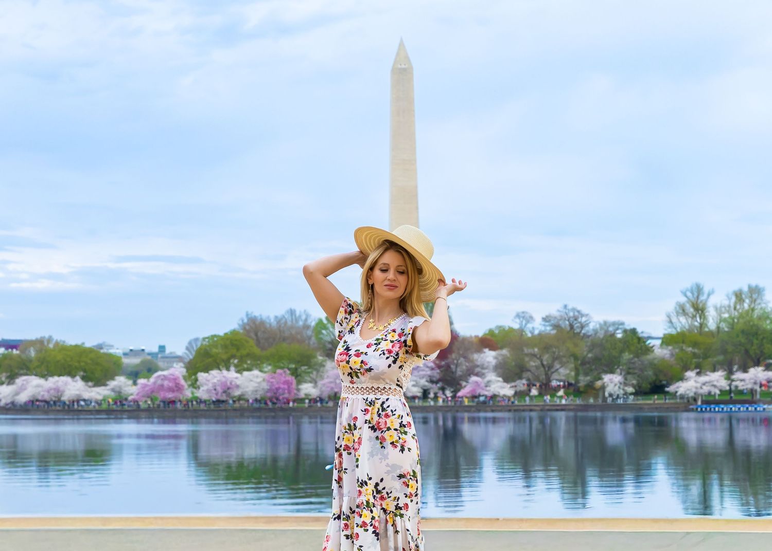 Woman in floral dress and straw hat poses by Tidal Basin with Washington Monument and cherry blossoms in background.
