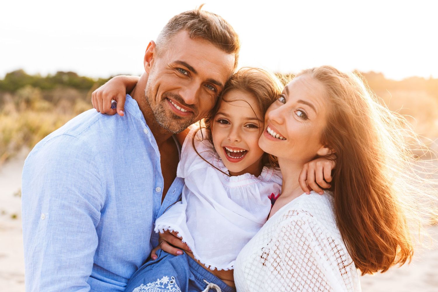 Happy family of three smiling together on a sunny beach, parents hugging their young daughter outdoors.