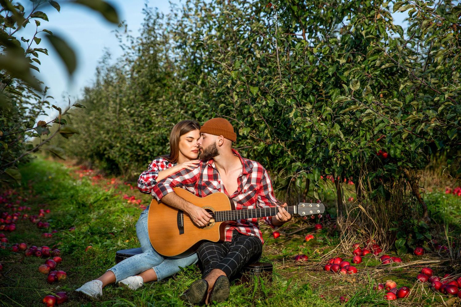 Couple sharing a romantic moment playing acoustic guitar in a lush apple orchard with red apples on the ground.