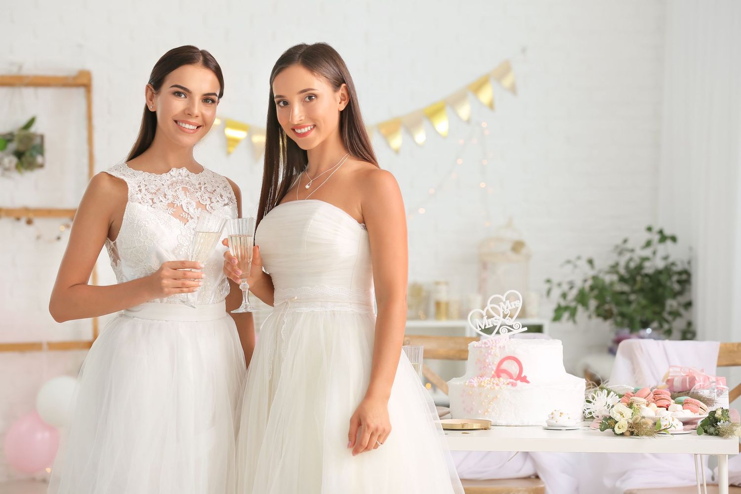 Two brides in white wedding dresses toasting with champagne glasses near a decorated wedding cake.