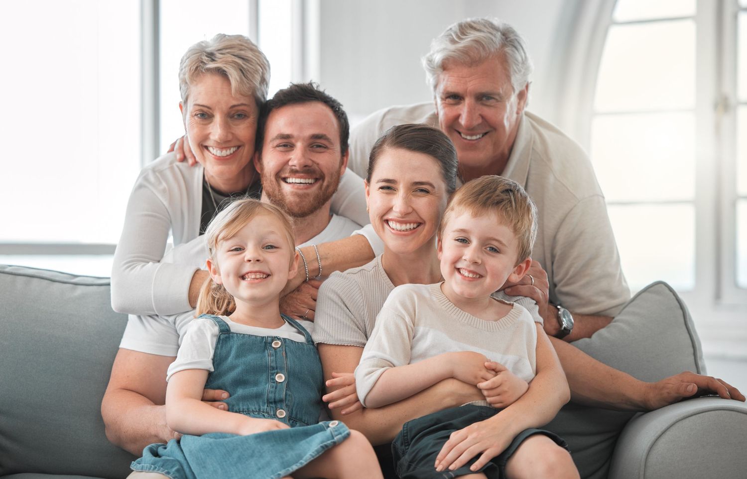 Happy multigenerational family of six smiling together on a couch in a bright living room.