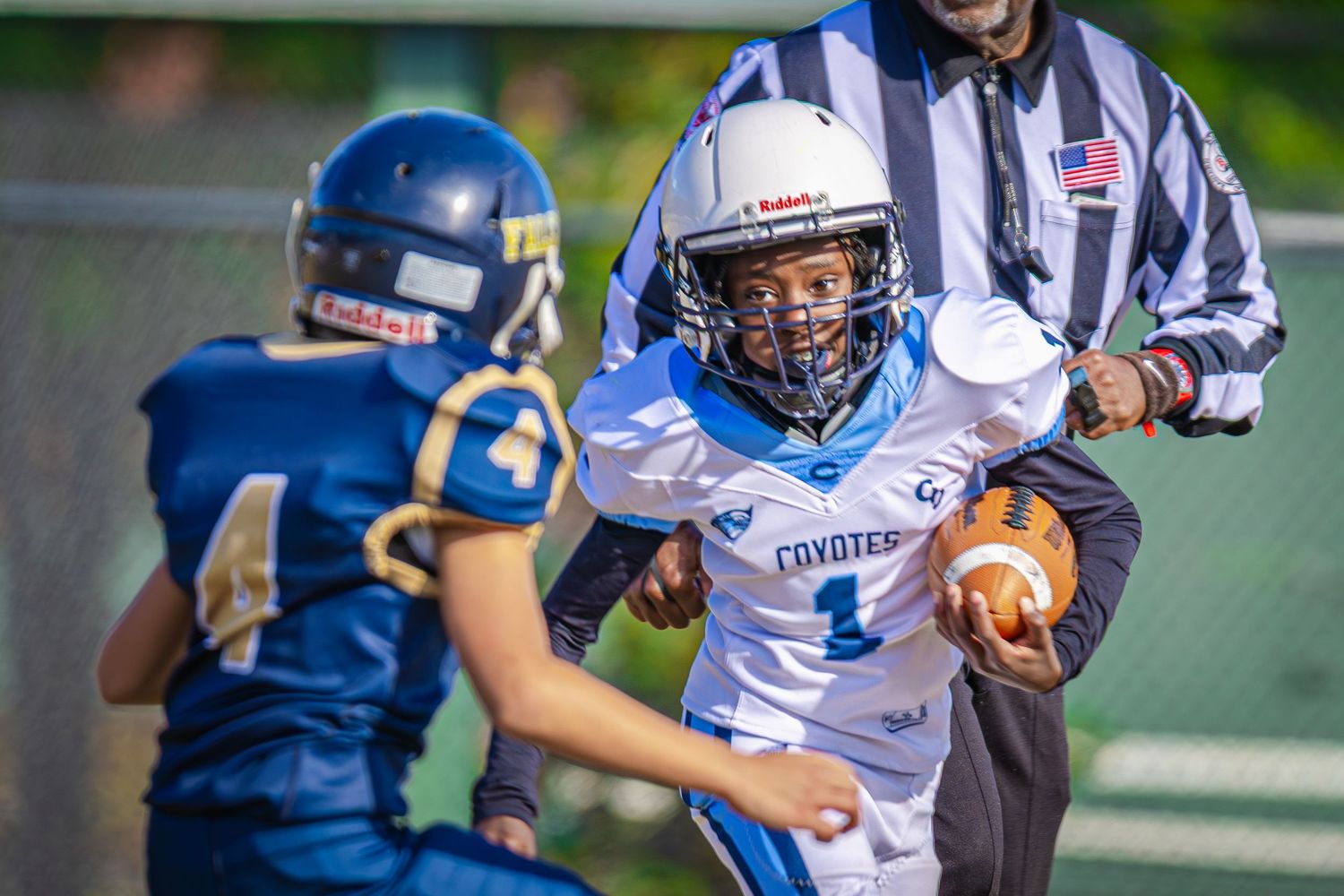 Youth football players in Coyotes uniforms compete during a game, with a referee watching nearby.