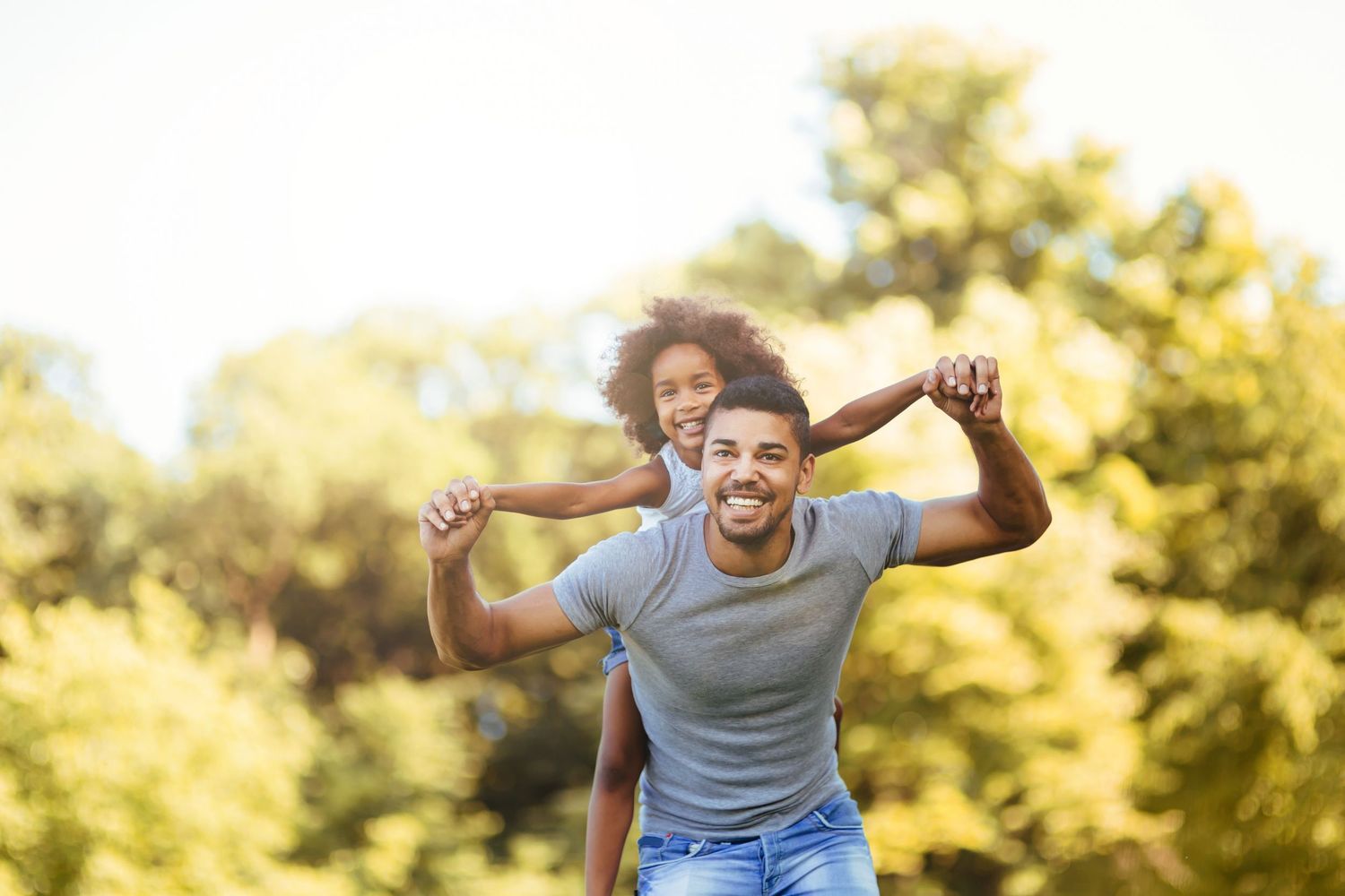 A smiling man carries a happy young girl on his shoulders outdoors in a sunny green park.