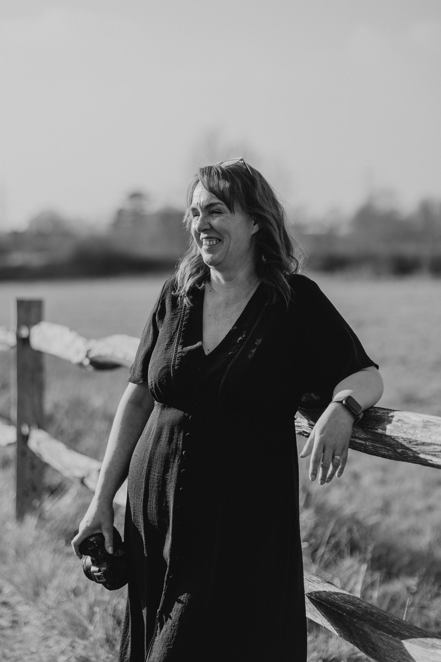 Black and white photo of a smiling woman in a dark dress leaning on a wooden fence in an open field.