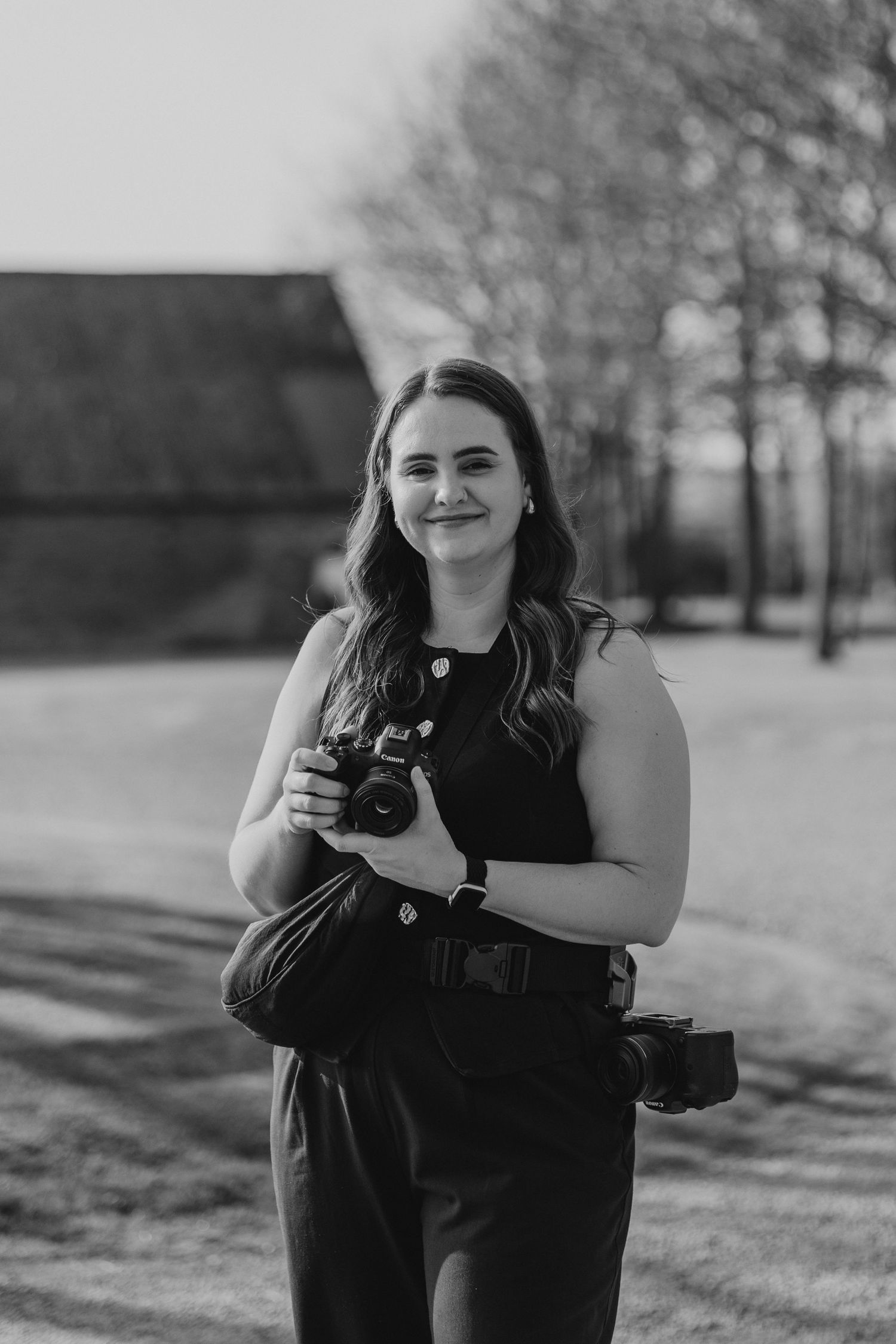 Black and white portrait of a smiling woman holding a camera outdoors near a barn.