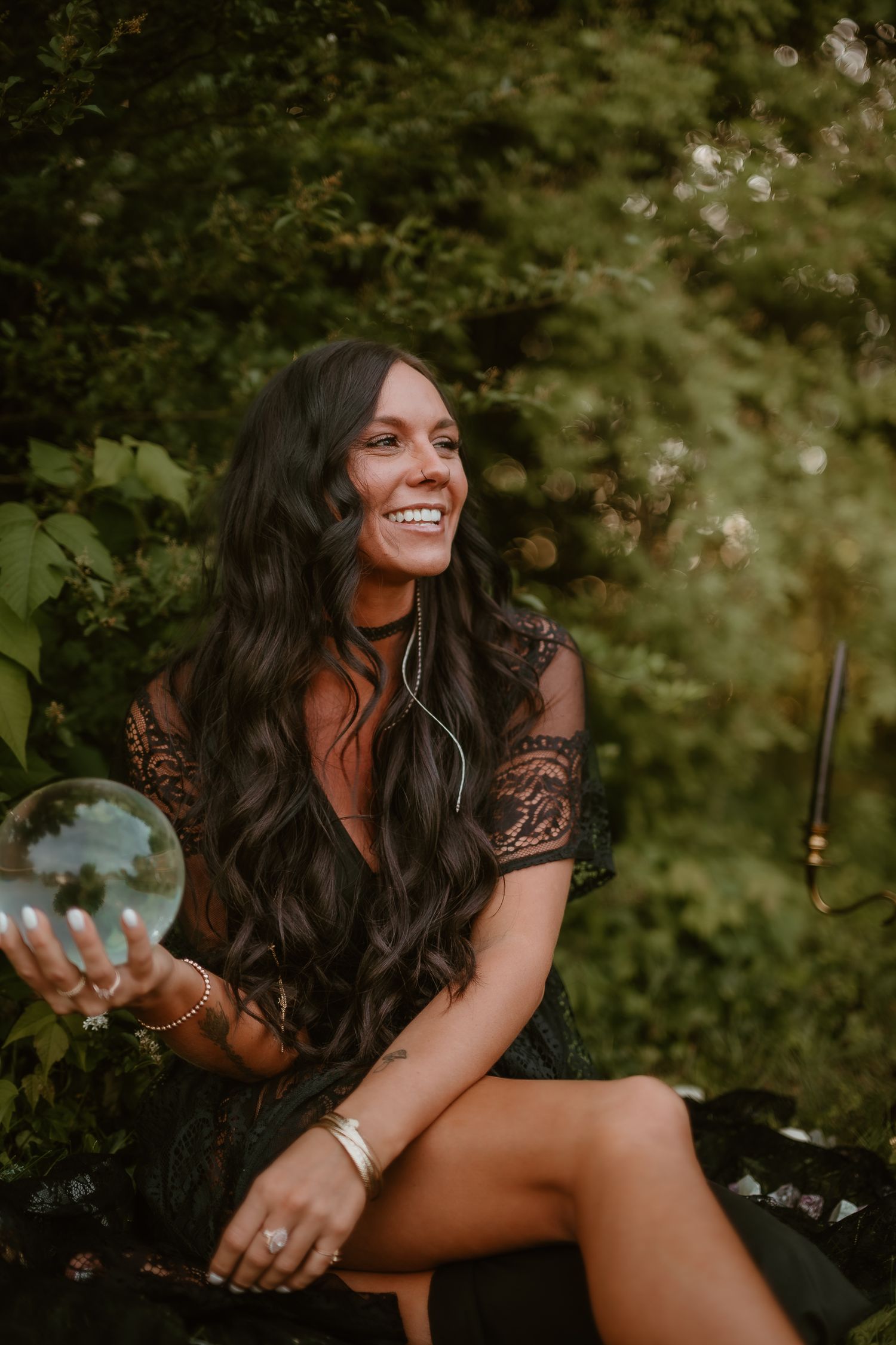 A woman in a dark lace dress sits outdoors holding a crystal ball with lush greenery in the background.