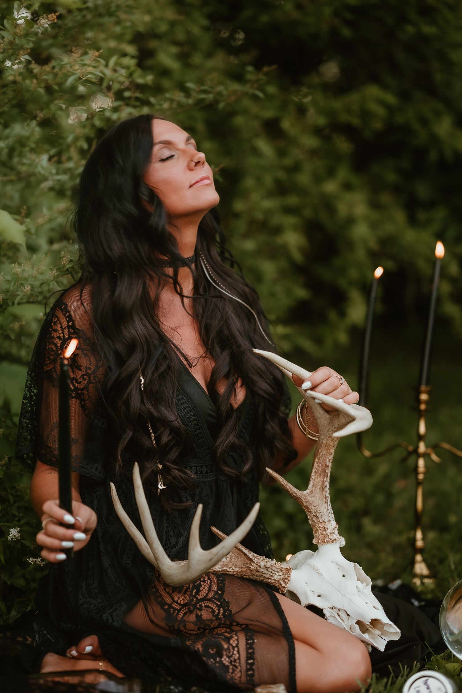 Woman in black lace dress holding deer skull while sitting in moody forest setting with glowing candles in background.