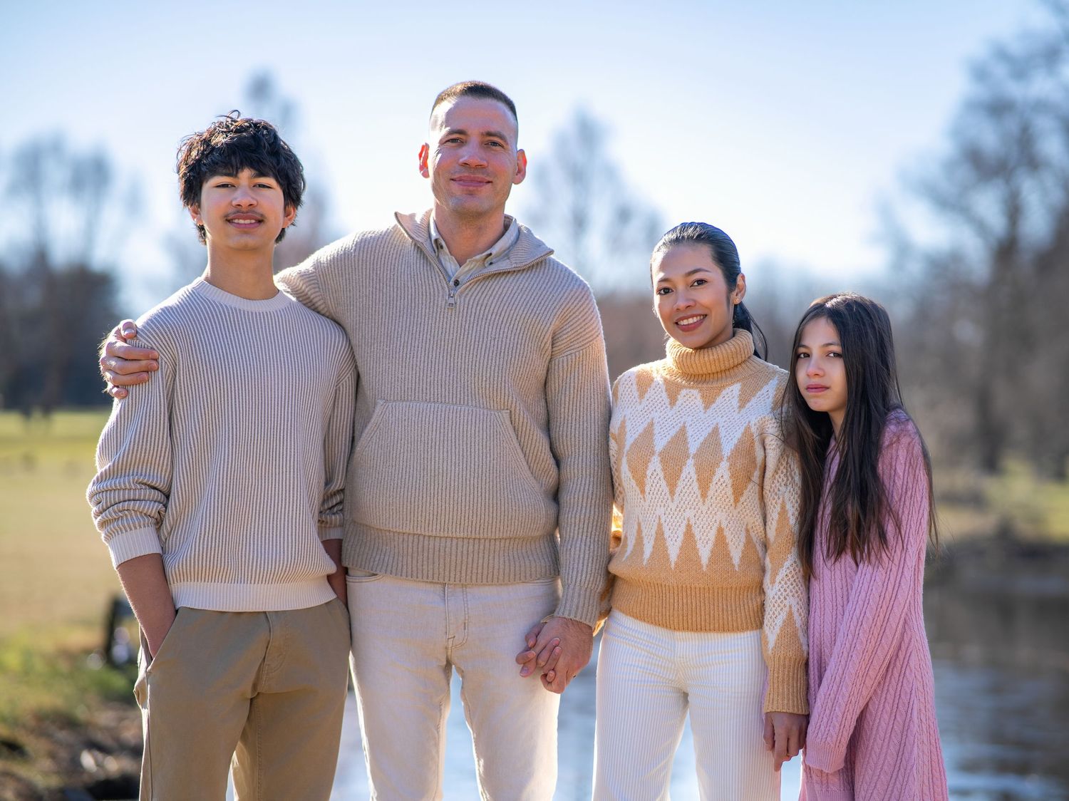 A group of four people in coordinated neutral and pastel sweaters pose together by a tranquil lake on a sunny day.