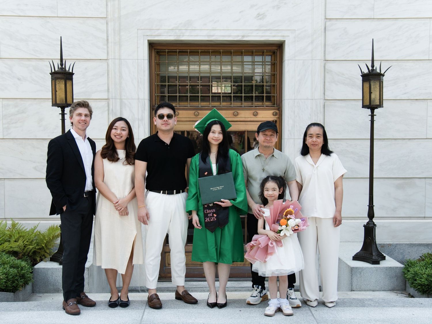 A group of seven people poses together outside a building with lantern light fixtures on a sunny day.