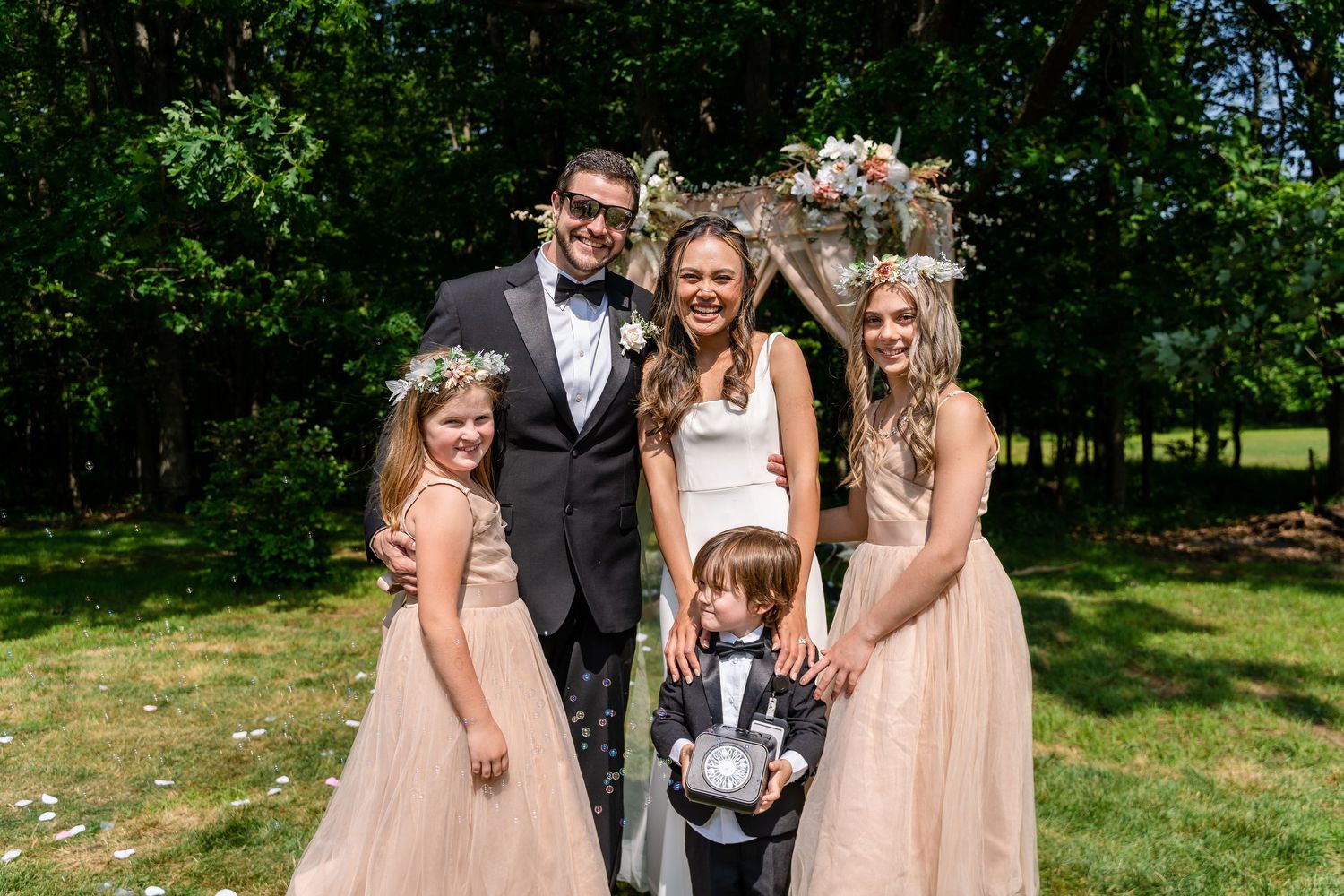 Wedding photoshoot service in Washington, D.C. Wedding party in formal attire poses together on sunny lawn with floral arch backdrop.