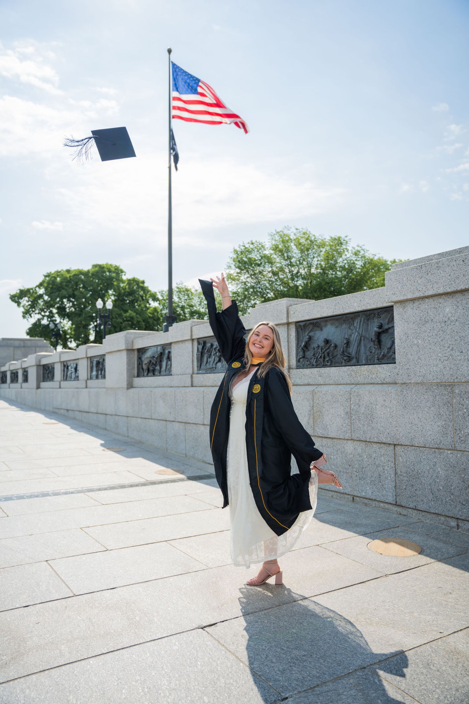 Smiling graduate in black gown tosses cap near American flag at WWII Memorial on a sunny day.