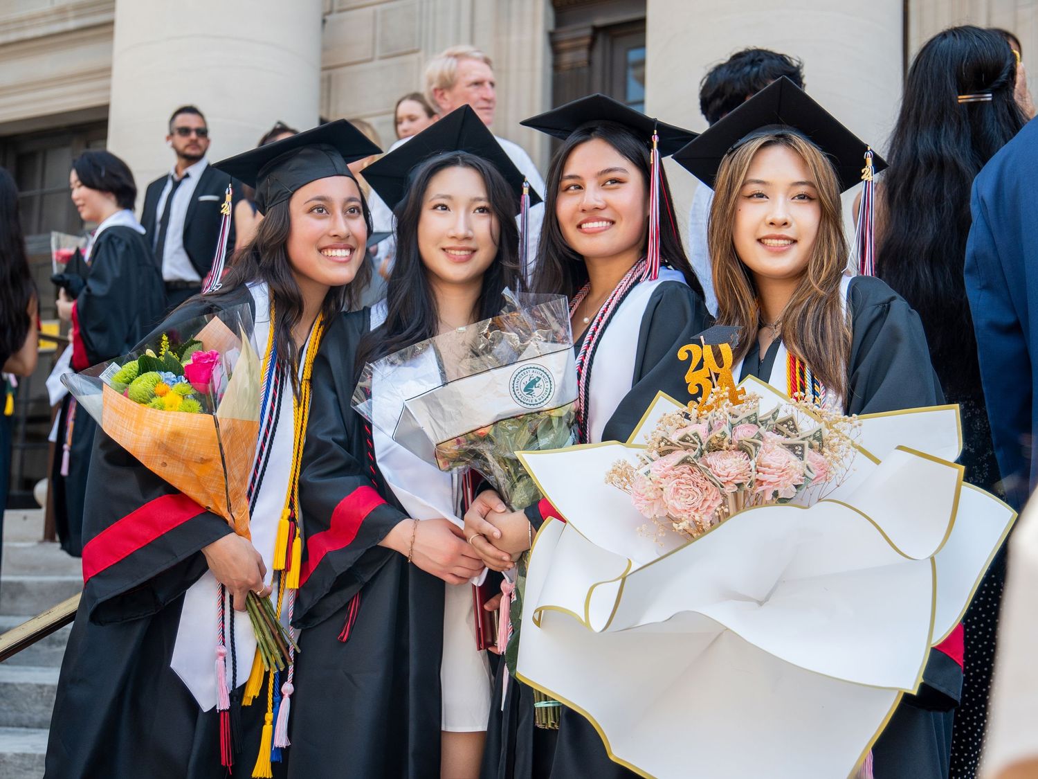 Four female graduates in caps and gowns smile holding flower bouquets at an outdoor graduation ceremony.