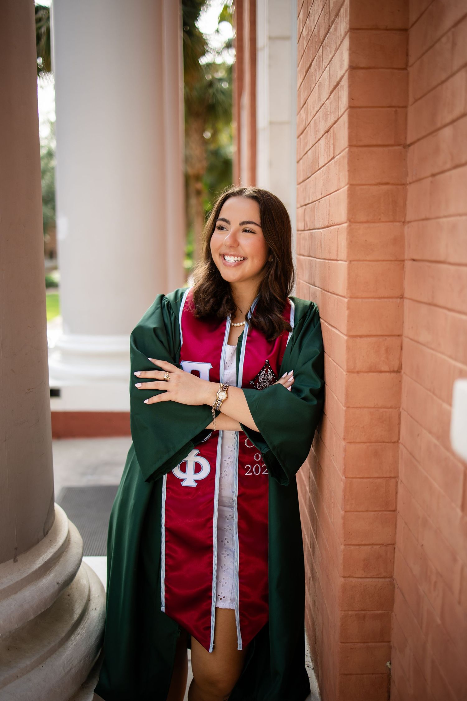 Graduate in green graduation gown and red stole poses in a series of portraits under pillared walkway.