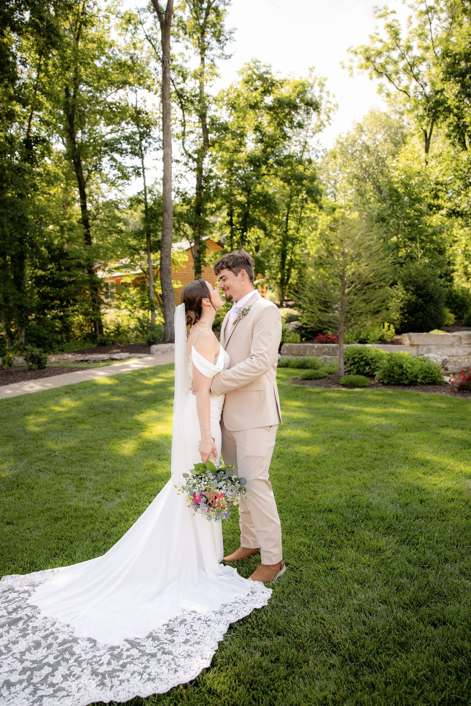 Bride and groom share a romantic moment on green lawn, surrounded by lush trees, bride in lace gown holding bouquet.