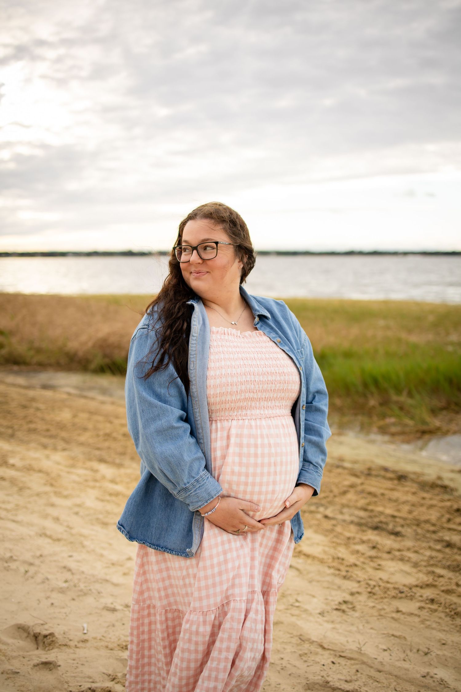 Woman in a pink gingham dress and denim jacket stands on a beach during overcast weather.
