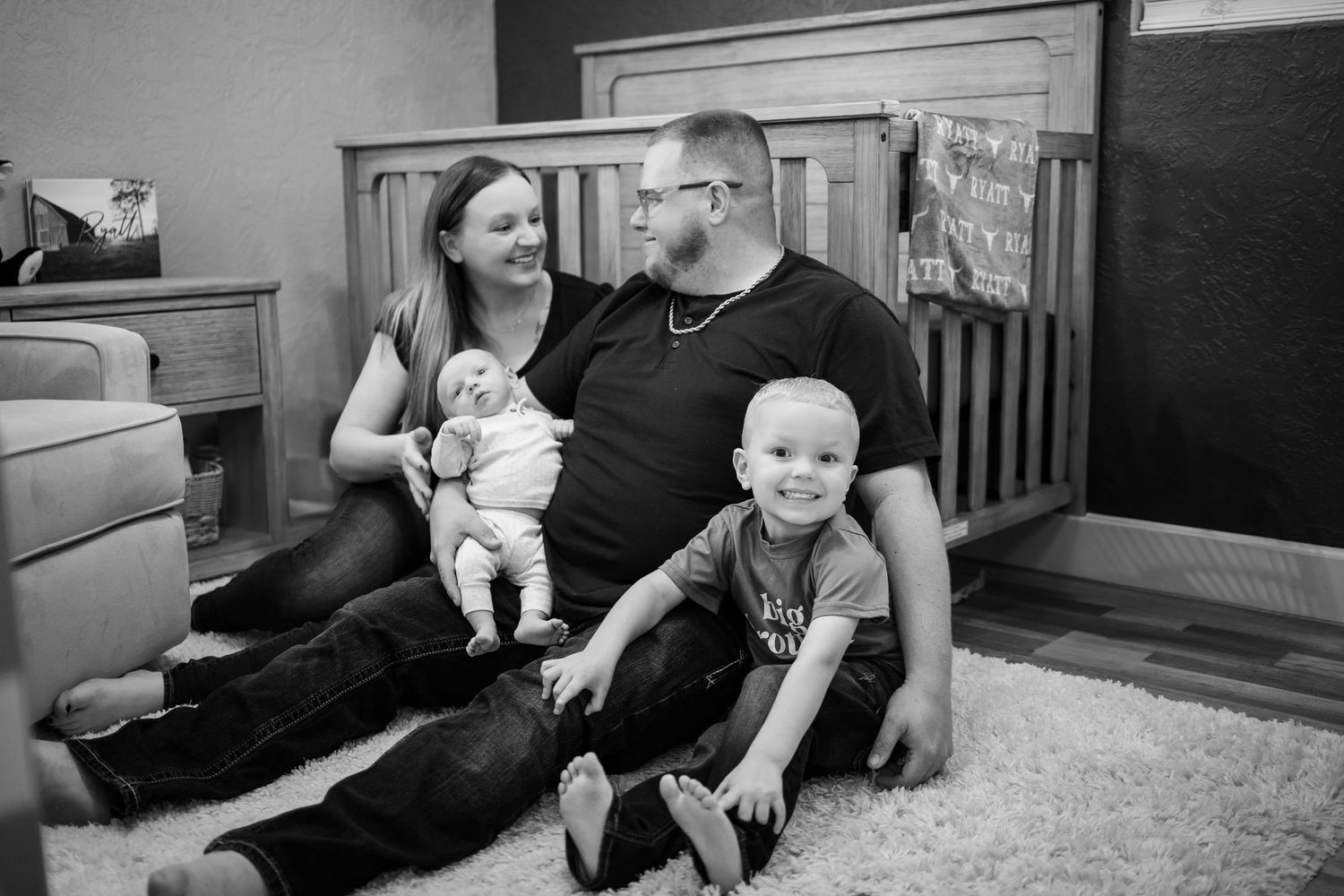 A family sits together on the floor of their home smiling in this intimate black and white portrait.