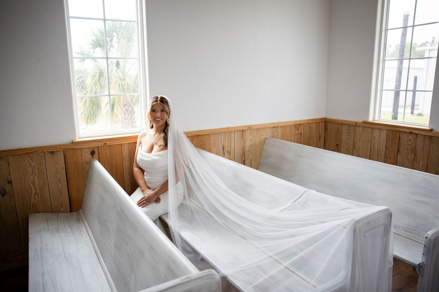 Bride in white gown and long veil poses between wooden church pews near sunlit windows on her wedding day.