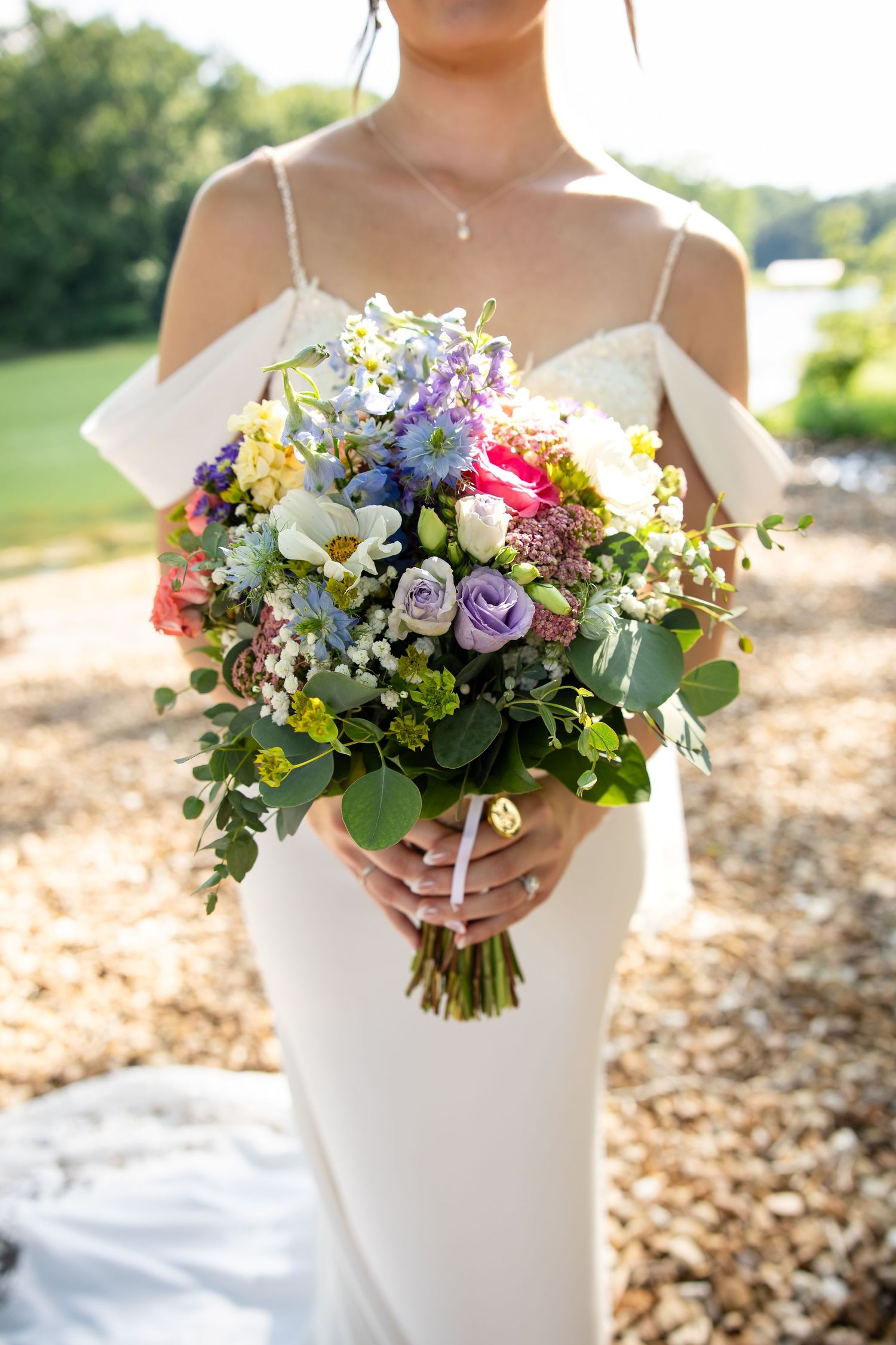 Bride in white off-shoulder gown holding a colorful wildflower wedding bouquet with roses and greenery outdoors.