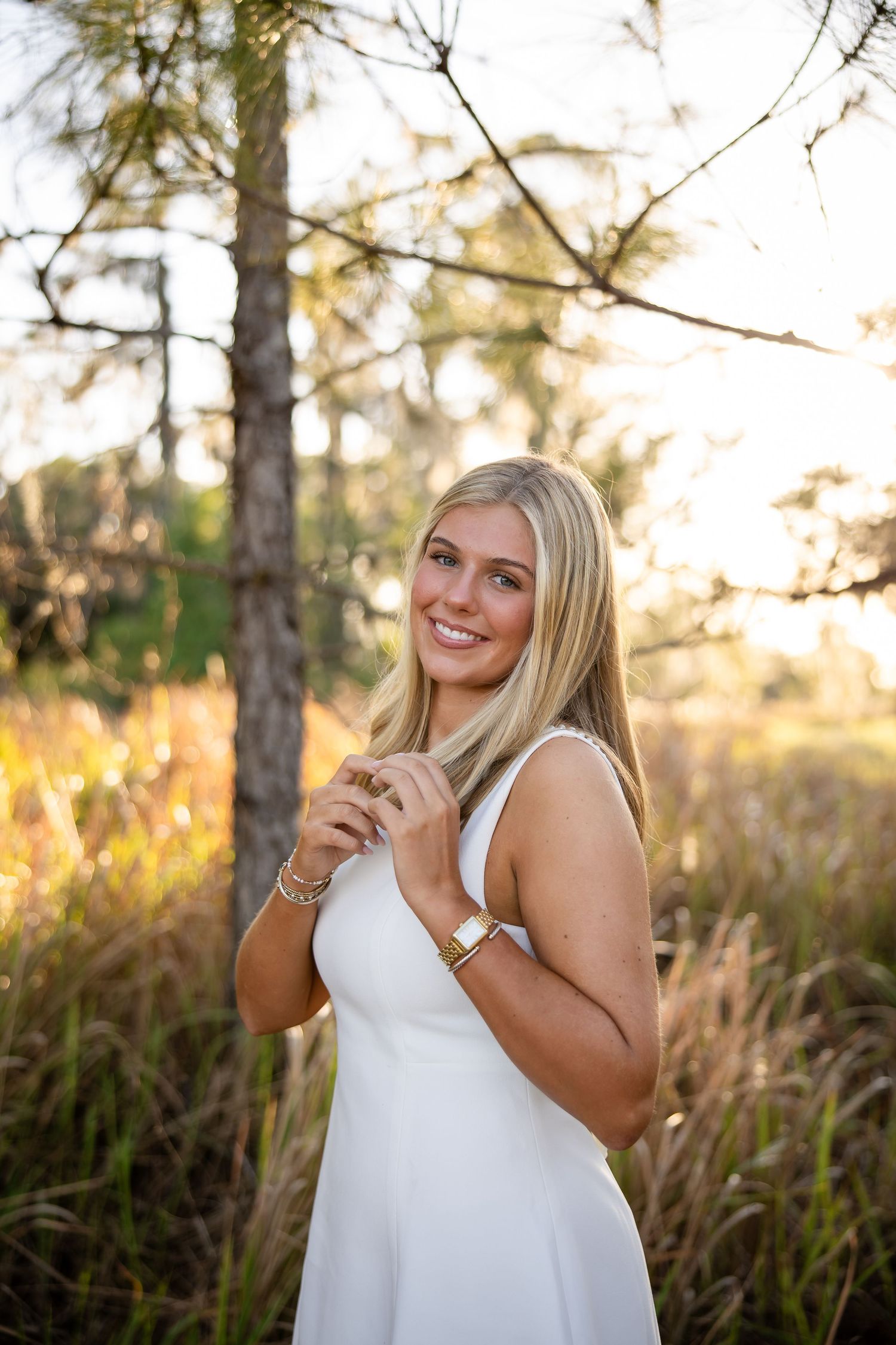 Young blonde woman in white dress smiling outdoors at golden hour with pine trees and tall grass background.