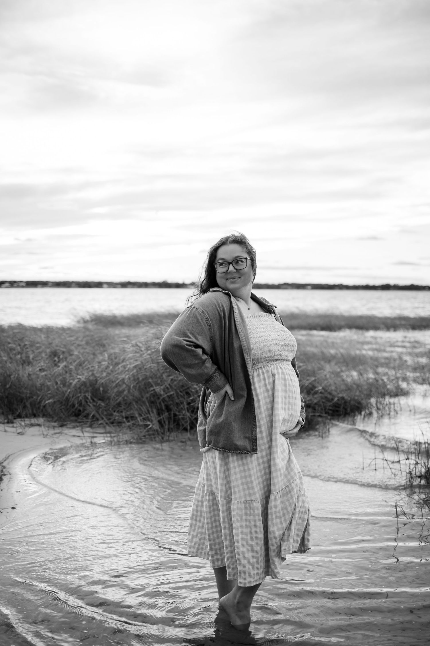 A black and white portrait of an expectant mother standing in coastal waters wearing a flowing dress.