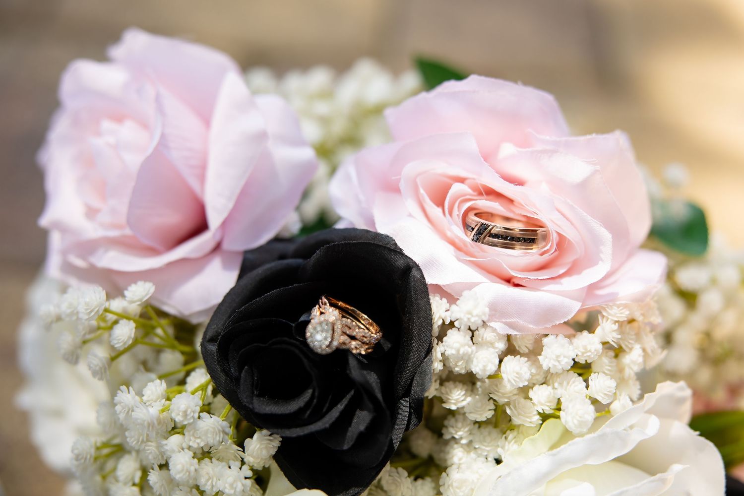 Wedding rings resting on pink and black roses with baby's breath in a bridal bouquet arrangement.