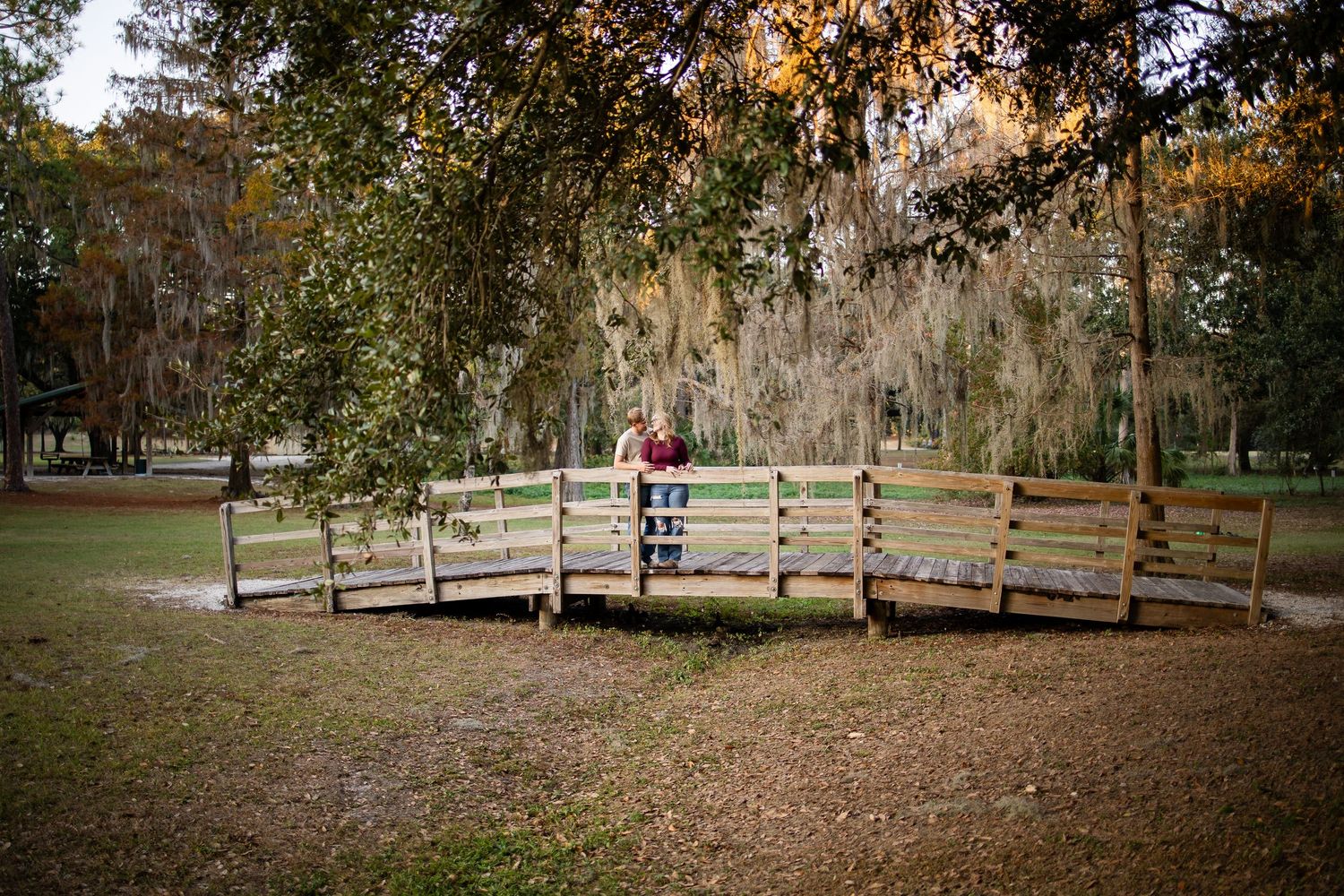 Couple standing on curved wooden bridge in park surrounded by oak trees.