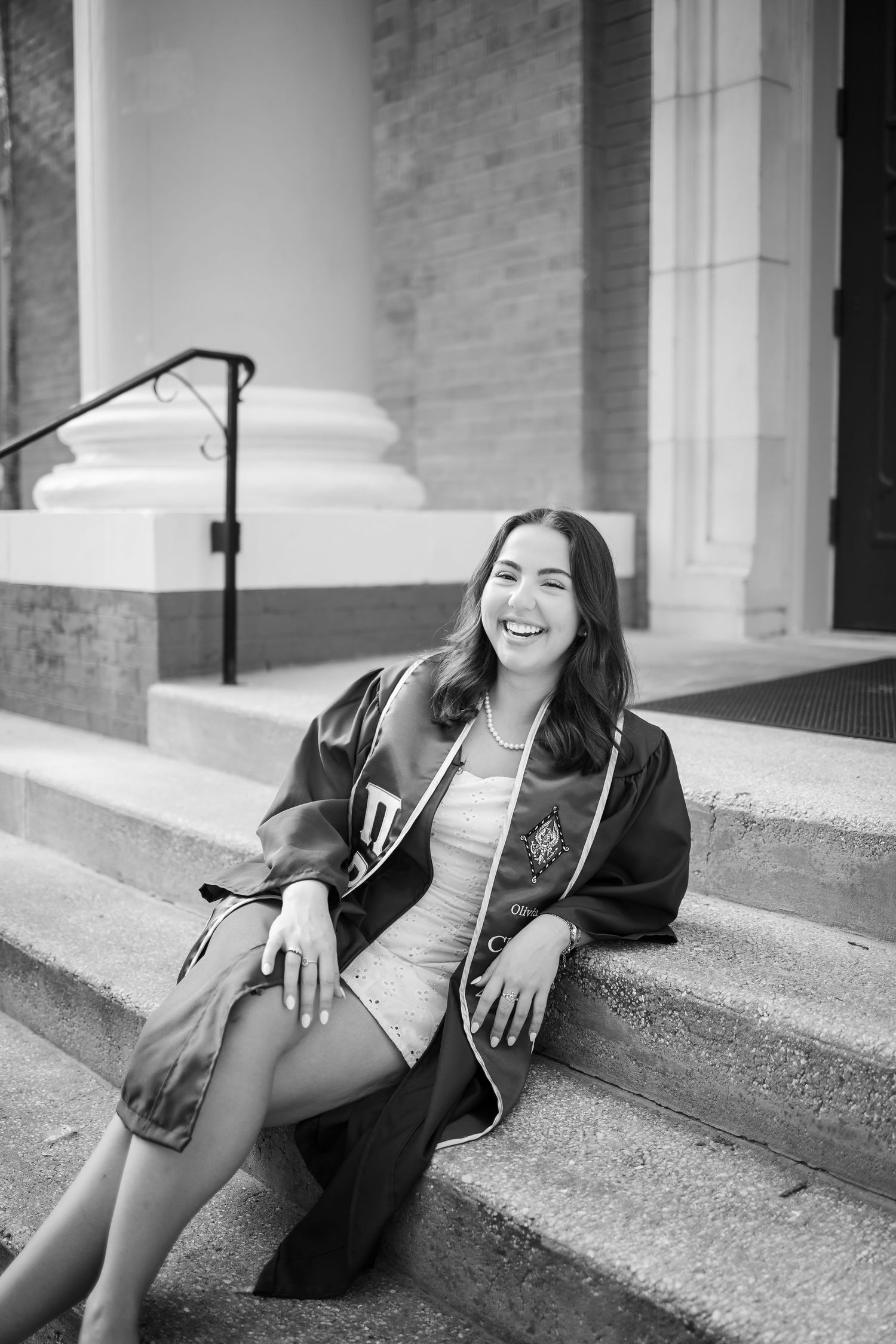 Black and white graduation photo of a smiling graduate in academic regalia sitting on stone steps of a building.