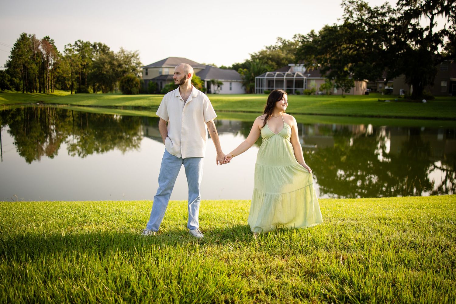 A romantic couple stands together on grass near a tranquil pond at sunset with mountains in the background.