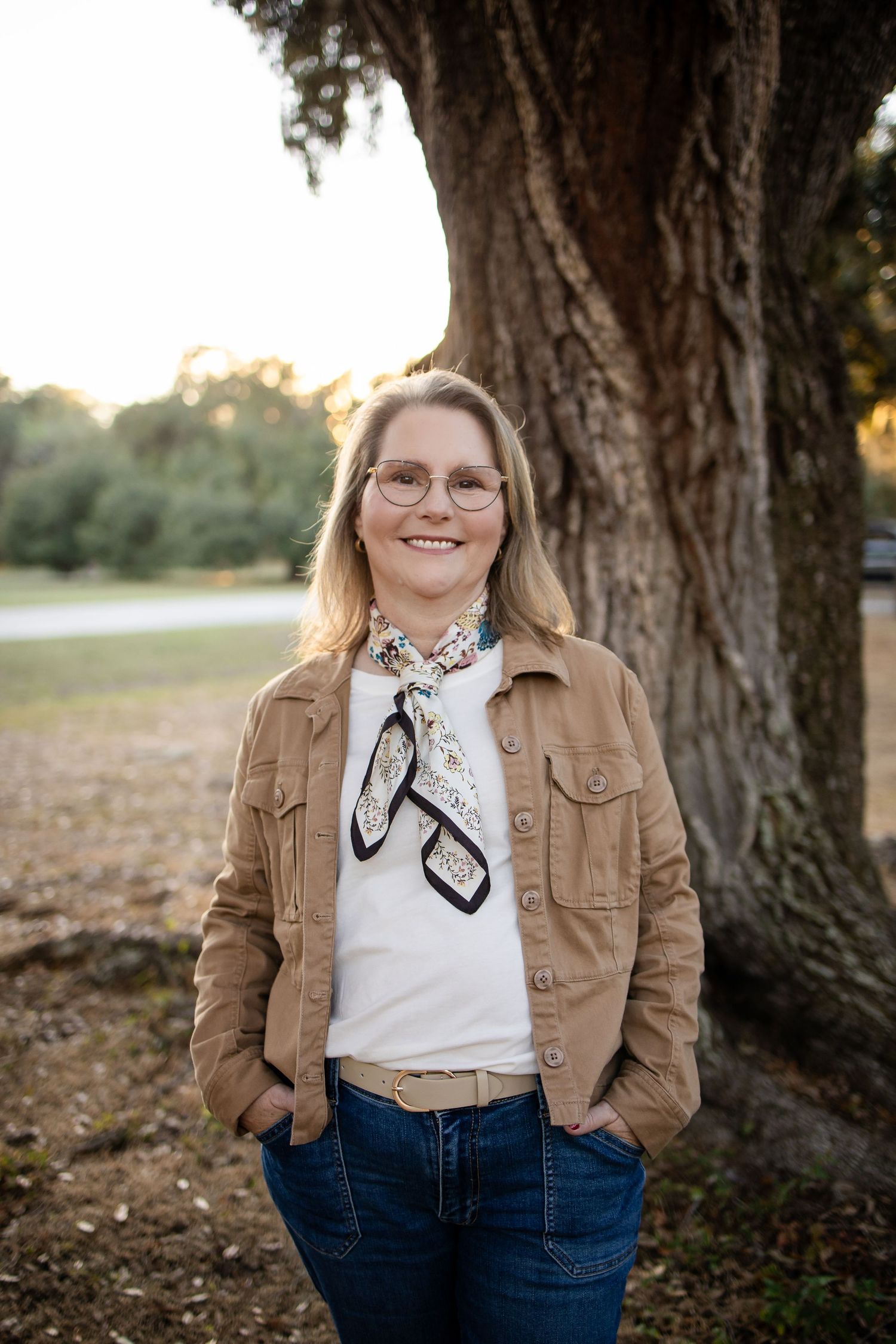Person in glasses wearing a tan jacket and silk scarf poses by a large tree trunk.