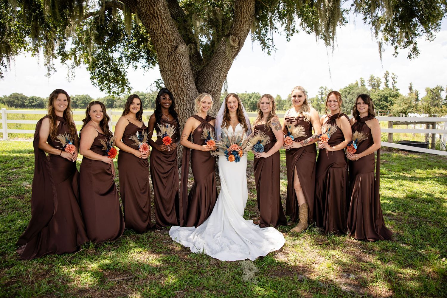 Bride in white gown with bridesmaids in brown dresses holding colorful bouquets under a large oak tree outdoors.
