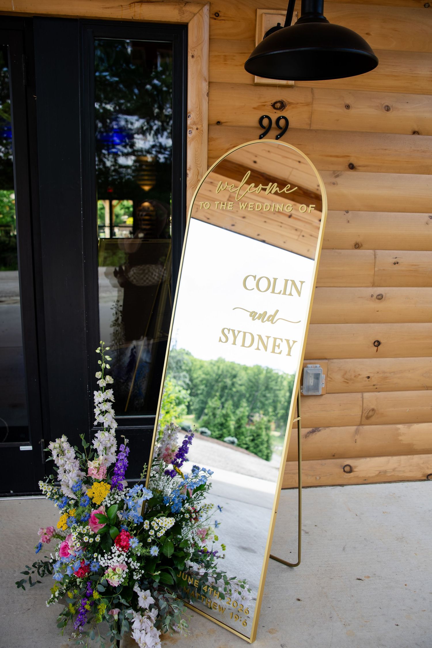Gold-framed welcome mirror sign for Colin and Sydney's wedding, adorned with colorful floral arrangement outside rustic wood venue.