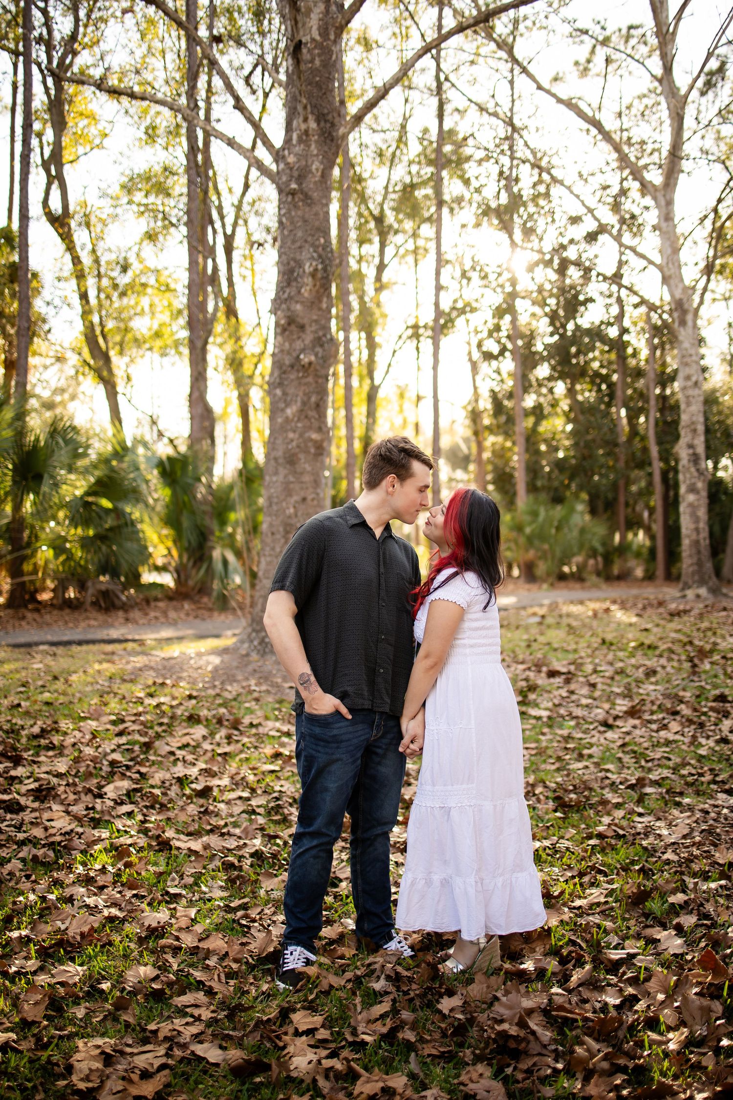 A couple shares a romantic moment together in a sunlit forest filled with fallen autumn leaves.