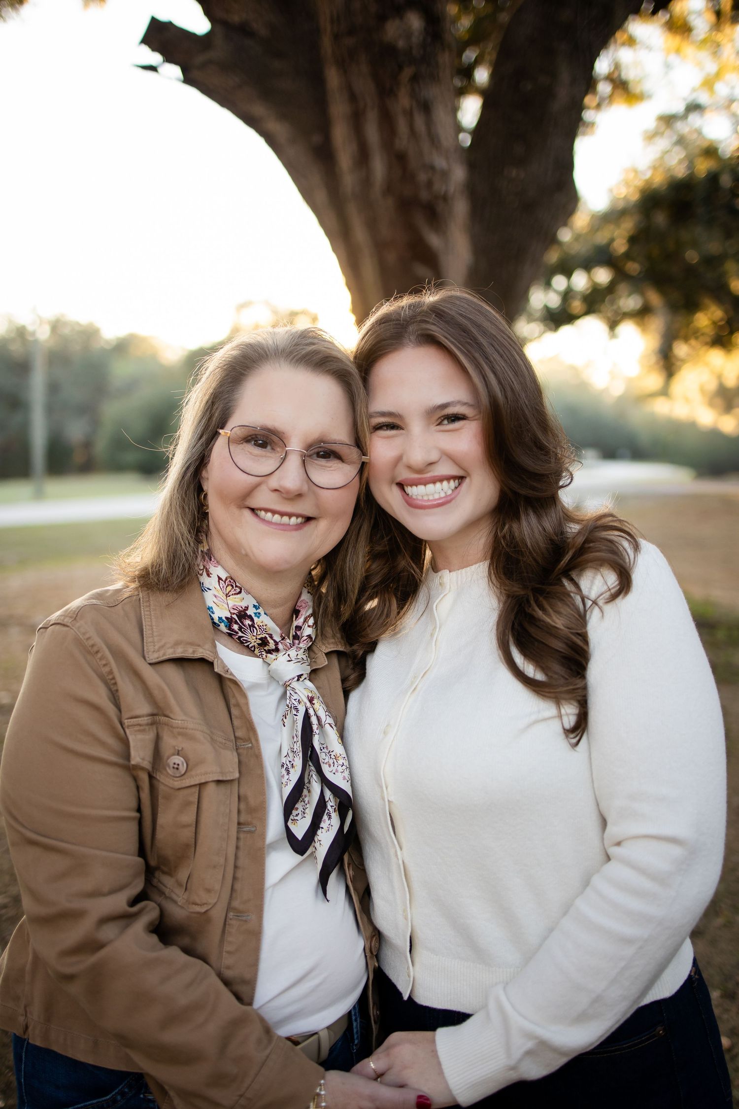 Two people embrace while wearing coordinating white sweaters and jackets under large tree.