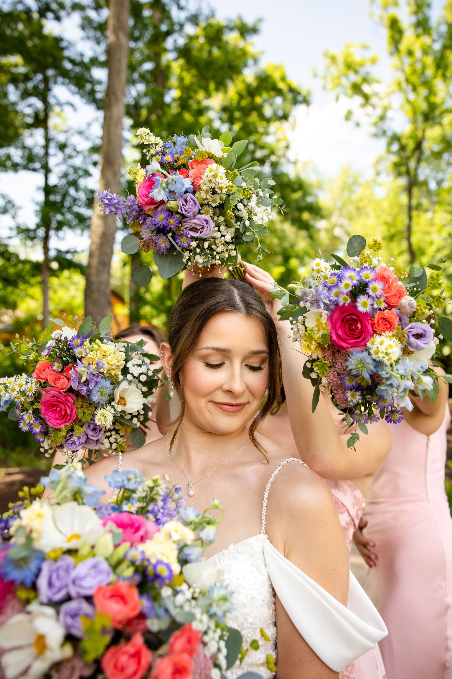 Bride surrounded by bridesmaids in pink dresses holding vibrant colorful wedding bouquets of roses and wildflowers outdoors.