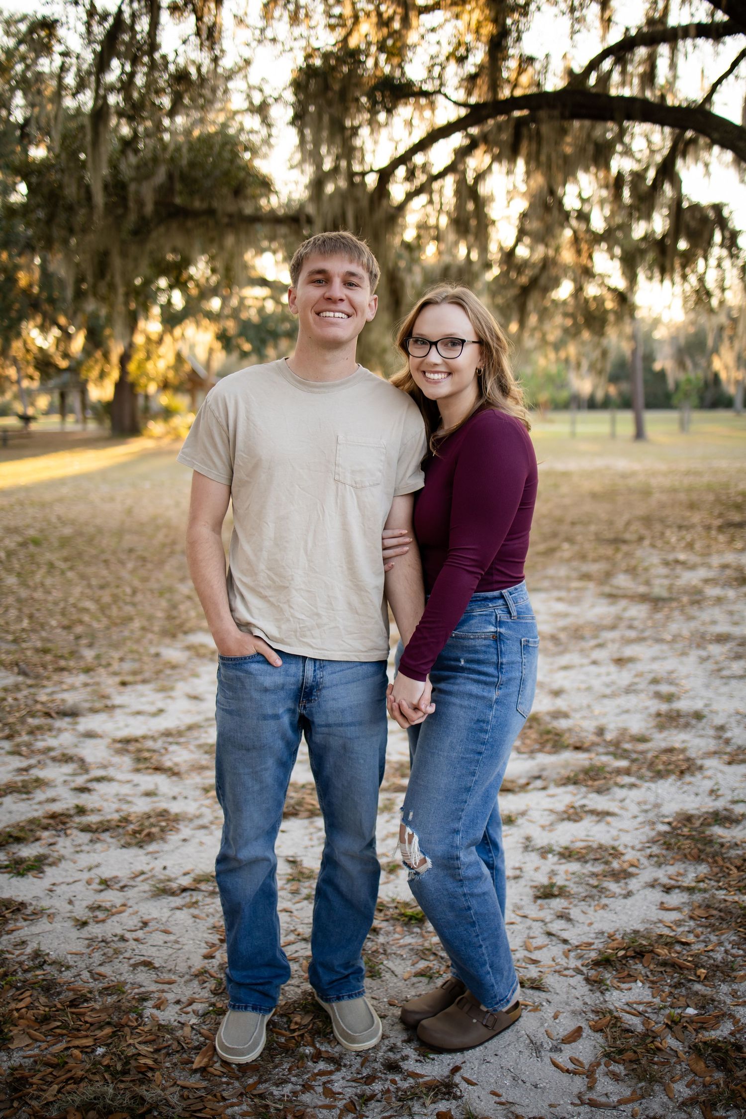 A couple in casual attire shares a tender moment at sunset in a park with Spanish moss draped trees.