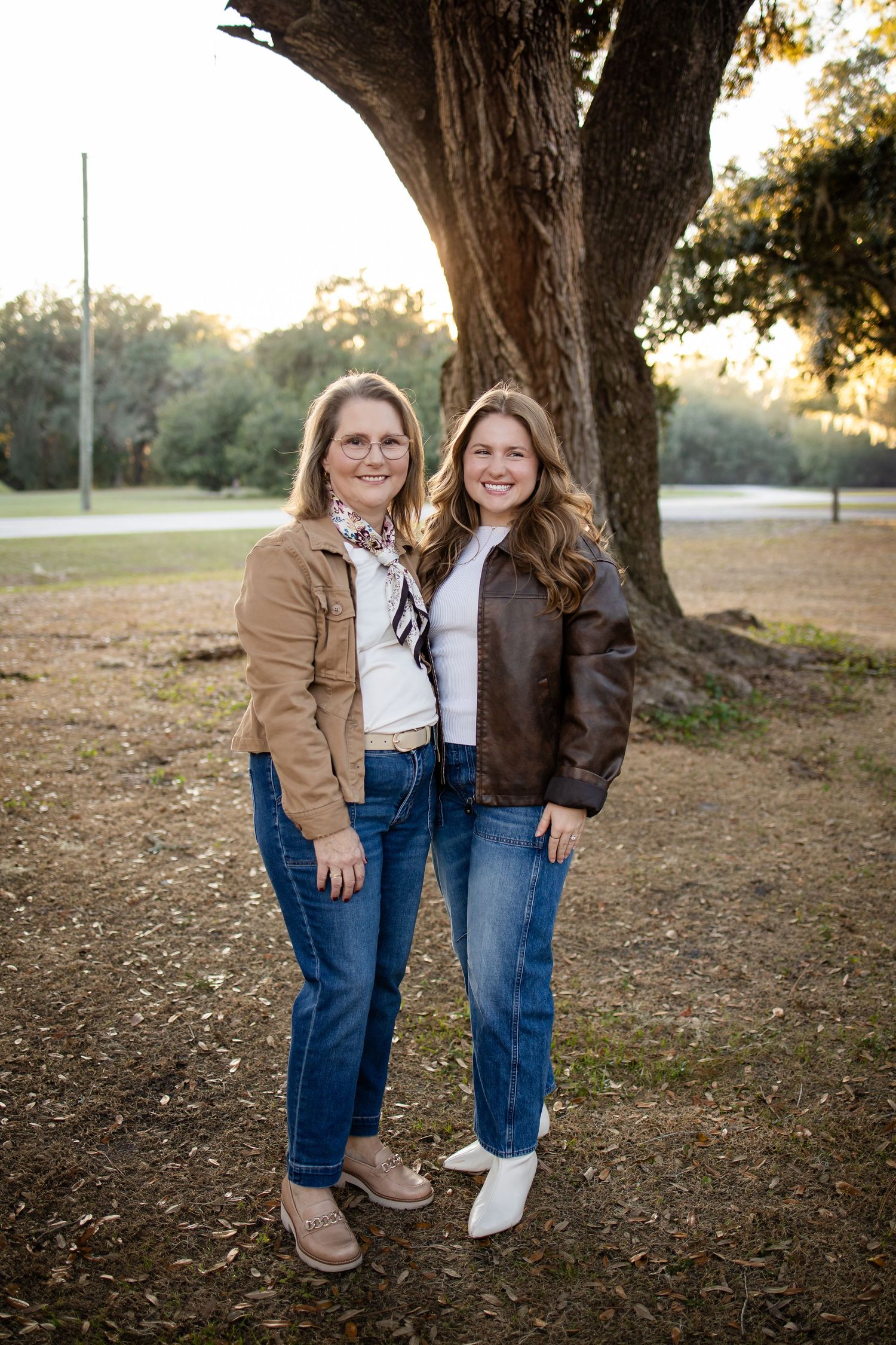 Two people in jackets and jeans stand together under a large tree during golden hour.