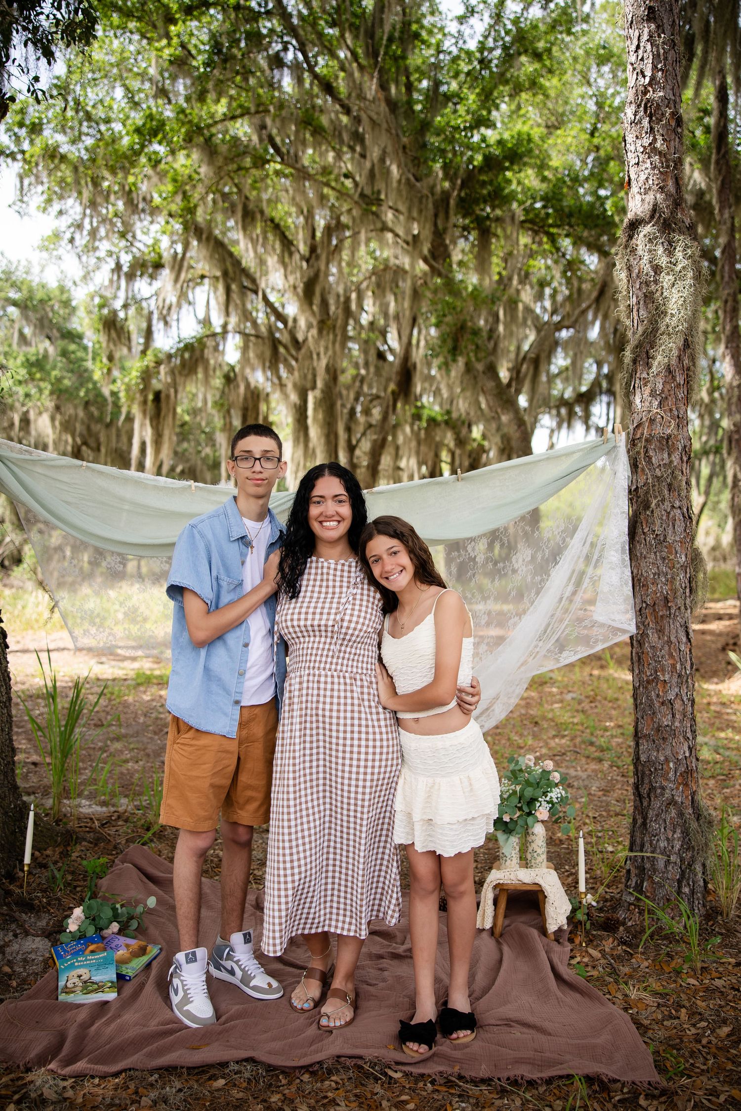 A woman and two teenagers pose outdoors under moss-draped trees with a sheer fabric backdrop on a brown blanket.