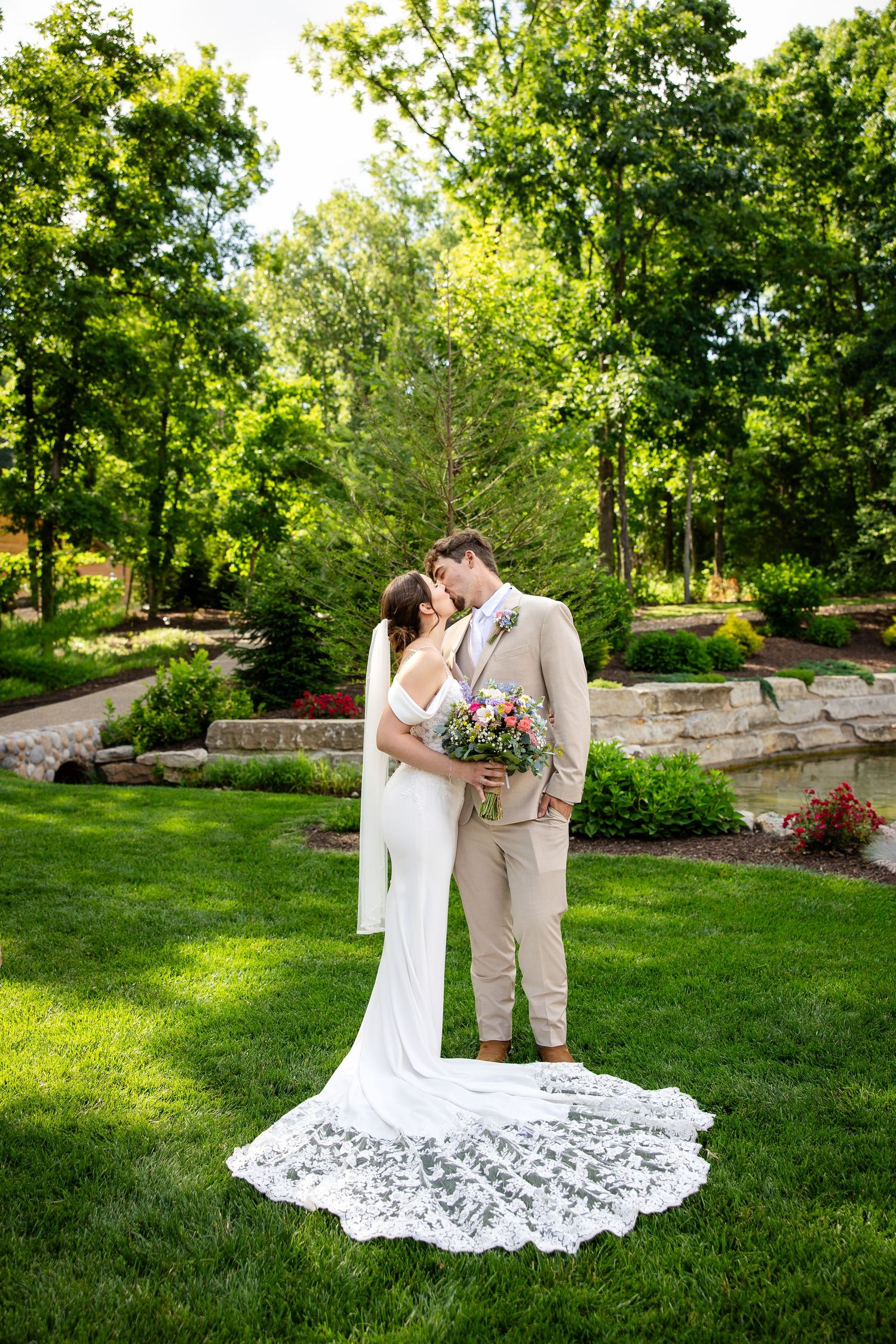 Bride and groom share a romantic kiss in a lush garden, bride wearing a white gown with lace veil train.