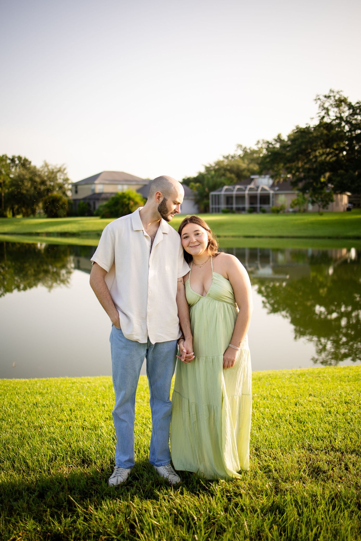 Pair in casual attire standing together on grass by a peaceful lake during golden hour.